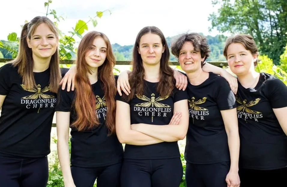 Fünf Frauen stehen nebeneinander im Freien, lächelnd, mit Arm in Arm. Sie tragen schwarze T-Shirts mit einem goldenen Libellen-Logo und der Aufschrift 'Dragonflies Cheer'.