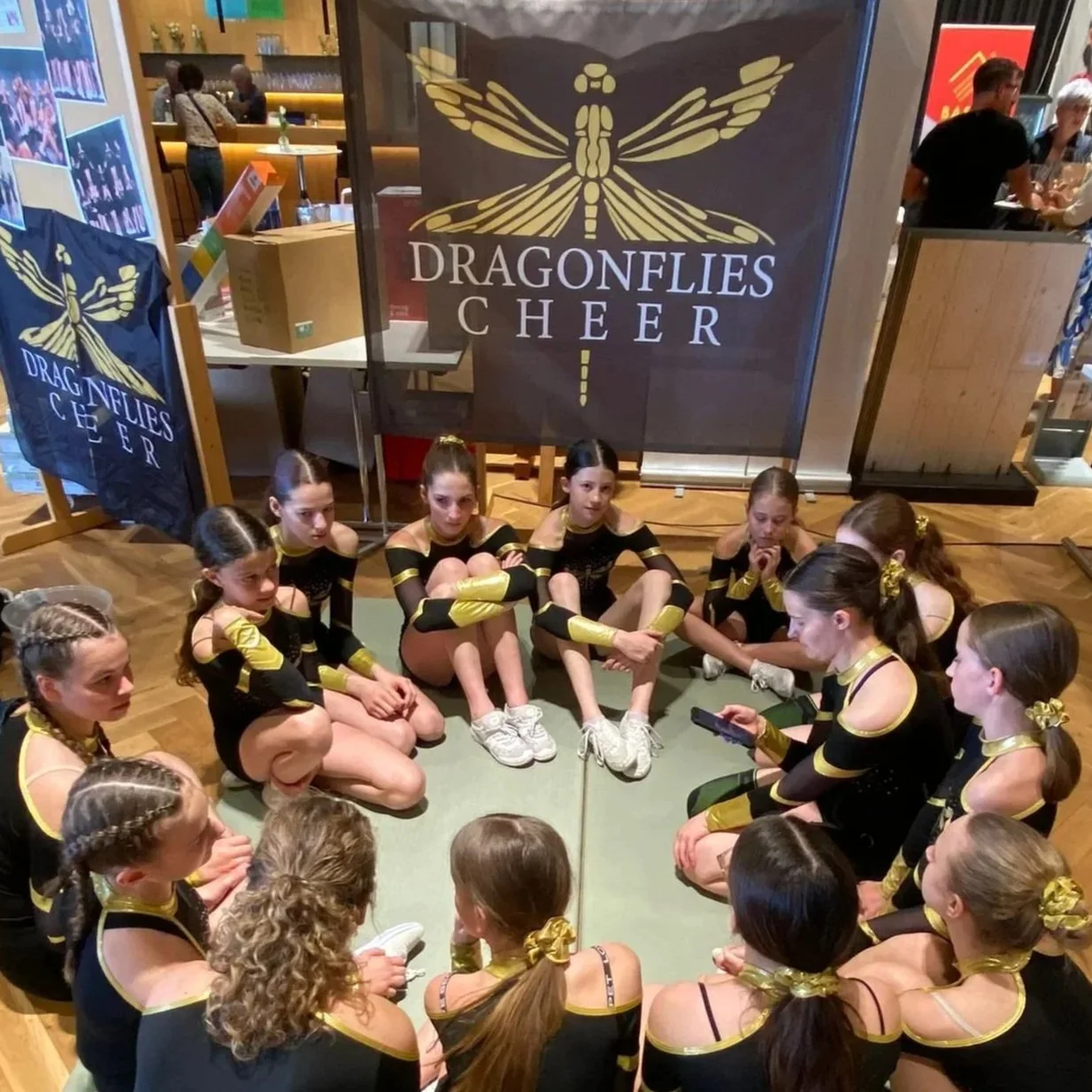 Gruppe von jungen Mädchen in schwarzen Turnanzügen mit goldenen Akzenten sitzen im Kreis auf dem Boden bei einer Veranstaltung. Im Hintergrund Tische, Leute und ein Banner mit einem Libellenbild.