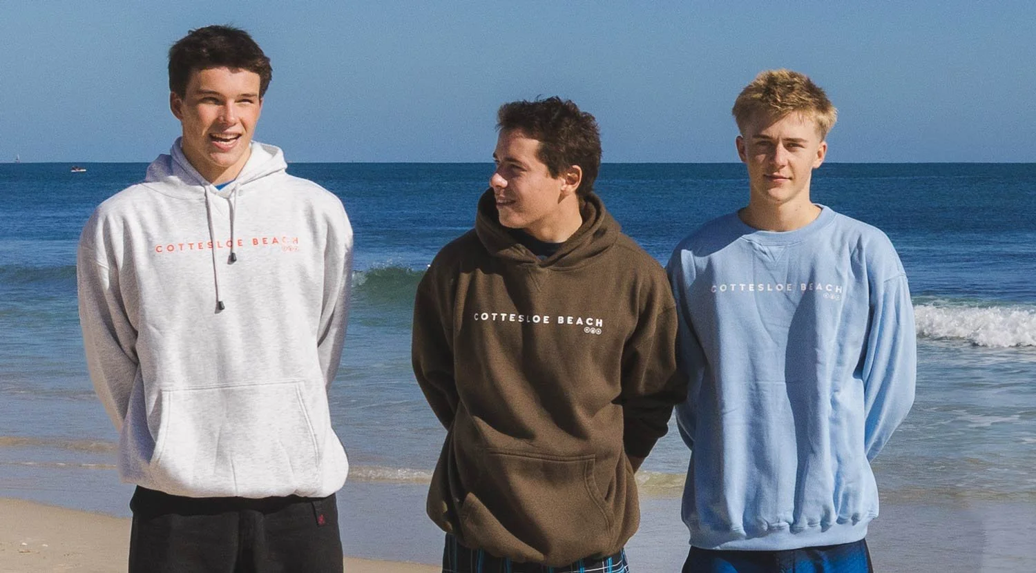 Three teenage boys standing on a beach with the ocean in the background, wearing Cottesloe Beach General Store hoodies.