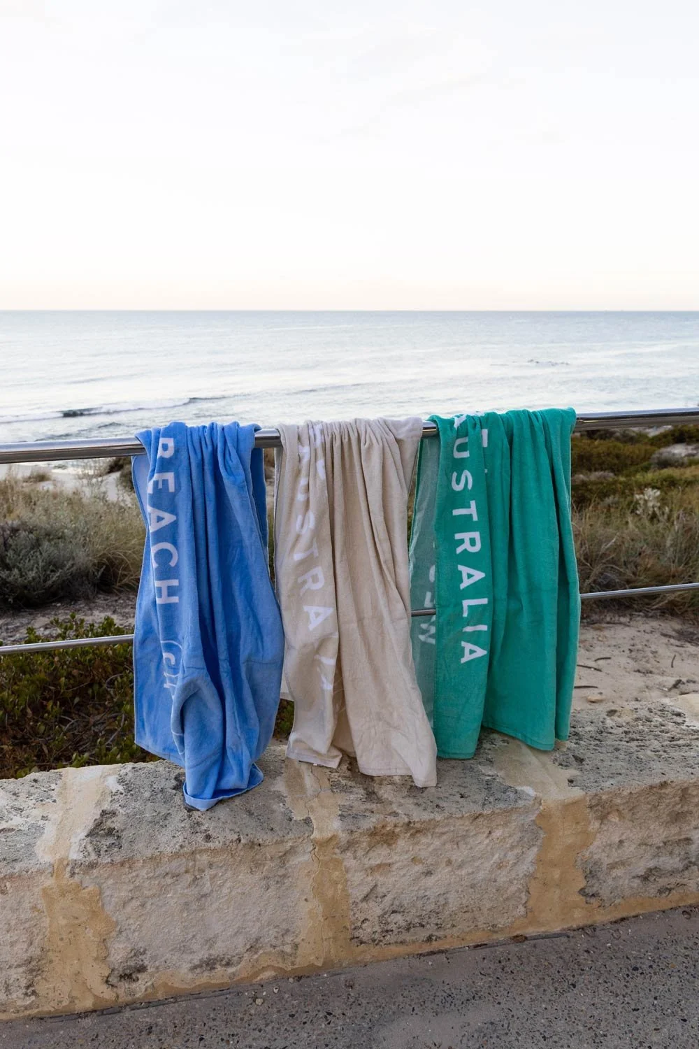 Three Cottesloe Beach General Store branded  beach towels hanging on a metal railing at Cottesloe beach with ocean and cloudy sky in the background.