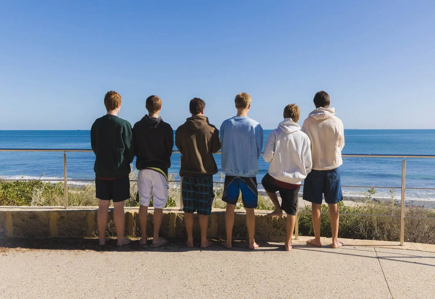 Six young men standing on a concrete pathway with their backs to the camera, looking out at the ocean on a clear day, wearing shorts and Cottesloe Beach General Store hoodies.