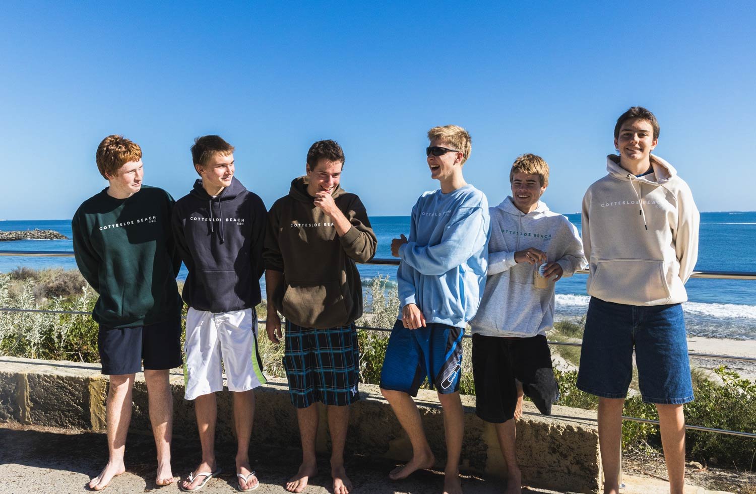 Six teenage boys standing barefoot on a beach, facing the camera and smiling, with the ocean and blue sky behind them. They are wearing shorts and Cottesloe Beach General Store hoodies enjoying a sunny day by the coast.