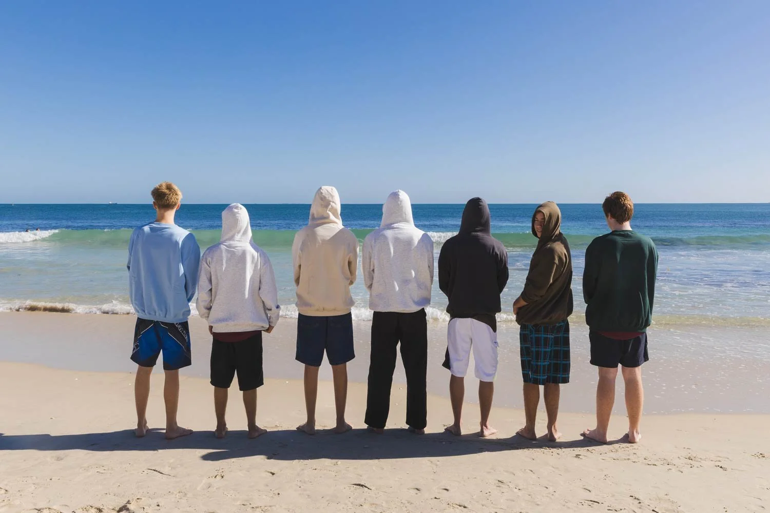Group of seven boys standing on the sandy beach, facing the ocean, wearing shorts and Cottesloe Beach General Store branded hoodies, during a sunny day with clear blue sky.