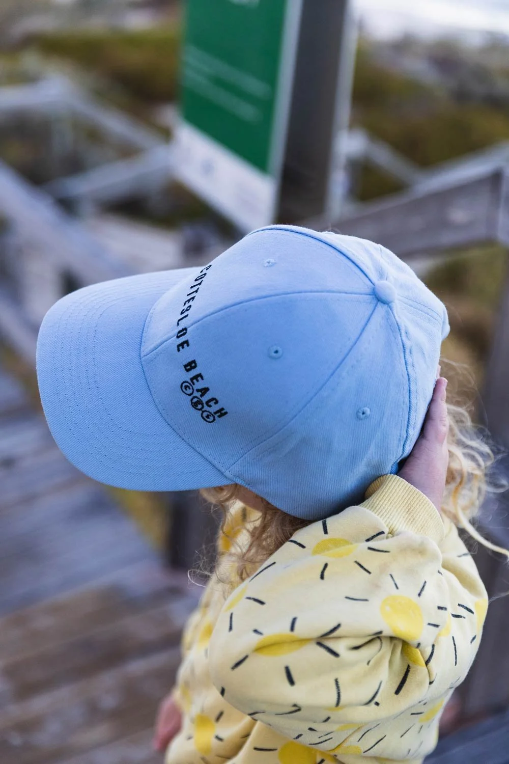 A child with curly blonde hair wearing a yellow sweatshirt and light blue Cottesloe Beach General Store branded cap, whilst on the stairs leading to Cottesloe Beach.