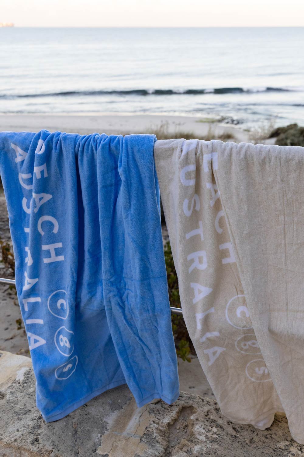 Two Cottesloe Beach General Store branded  beach towels hanging on a metal railing at Cottesloe beach with ocean in the background at sunset.