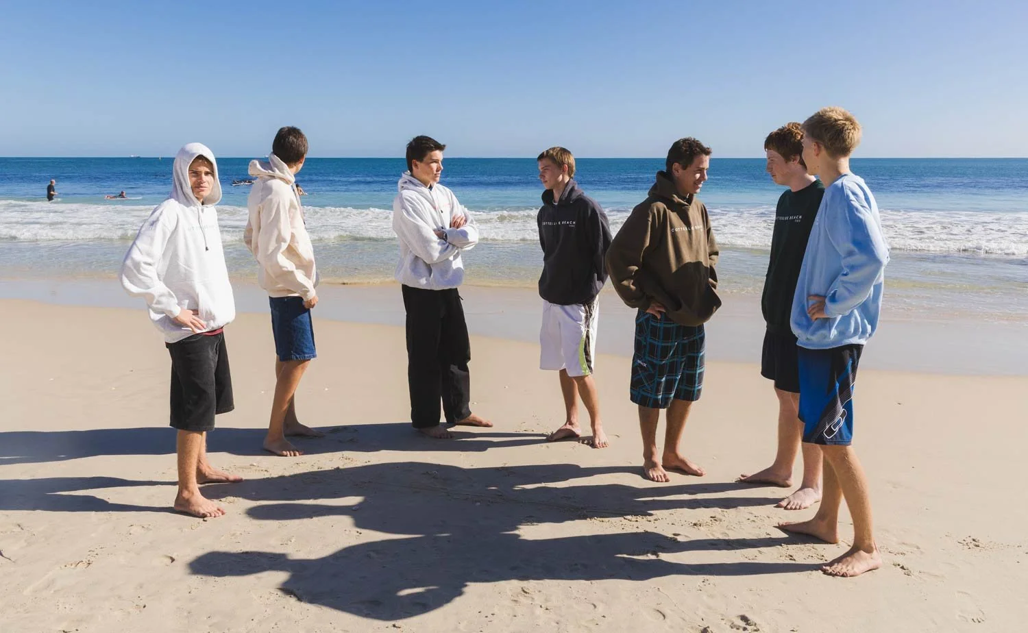 Group of young men standing on Cottesloe beach, facing each other wearing shorts and Cottesloe Beach General Store hoodies, with the ocean in the background.