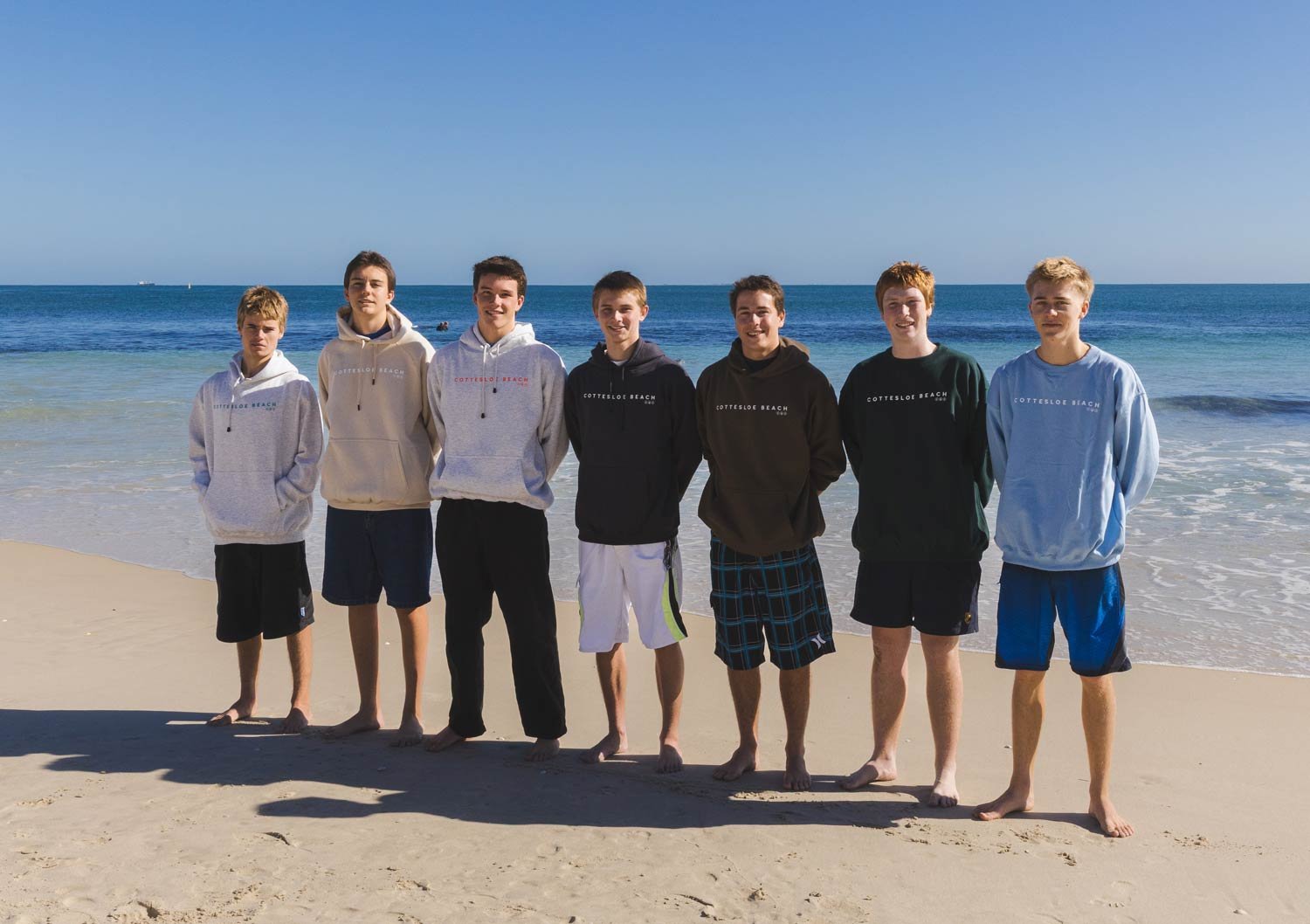 Seven teenage boys standing side by side on a sandy beach with the ocean and a blue sky in the background wearing shorts and Cottesloe Beach General Store branded hoodies.