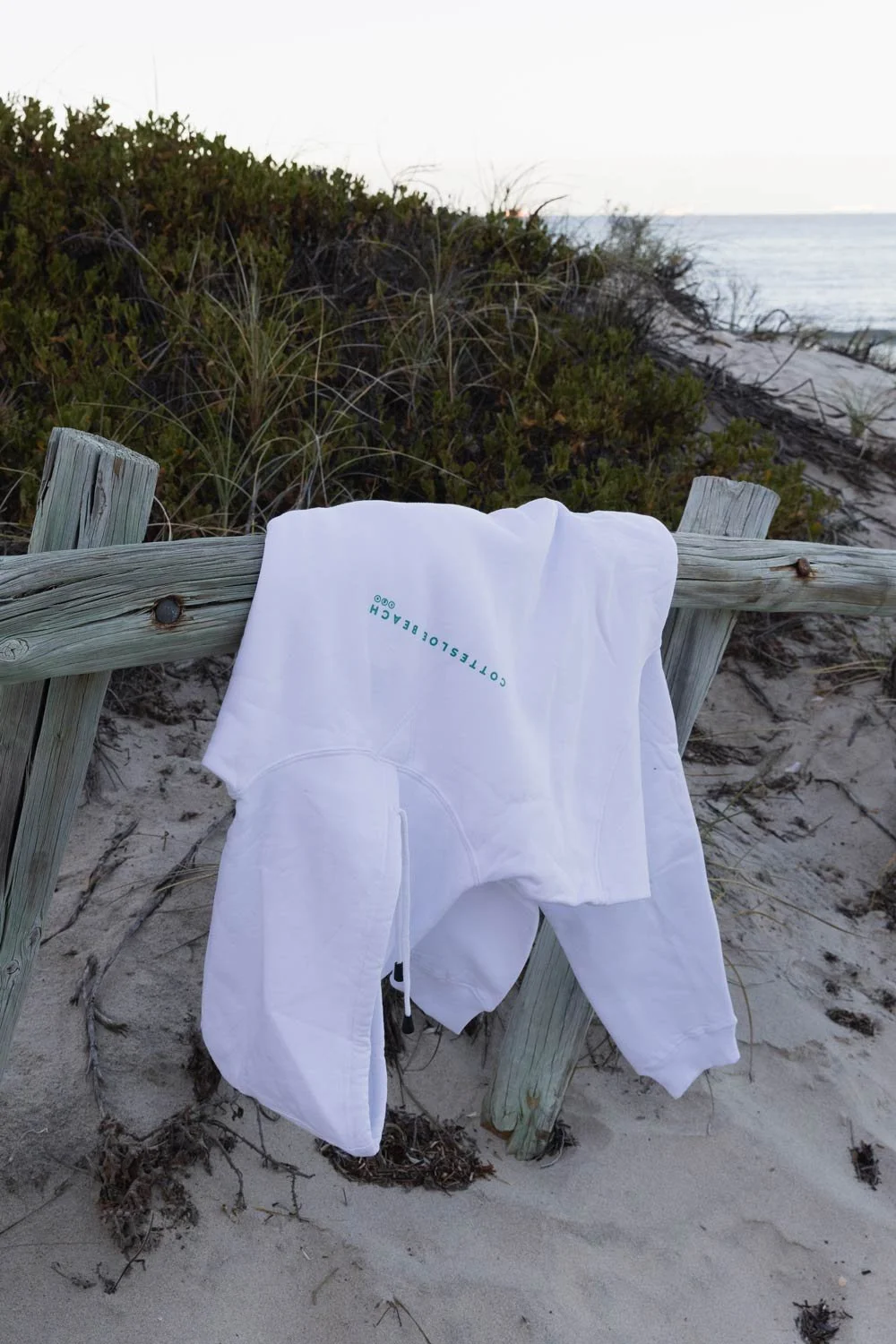 White Cottesloe Beach General Store branded hoodie draped over a weathered wooden fence on a sandy beach with dune grass and ocean in the background.