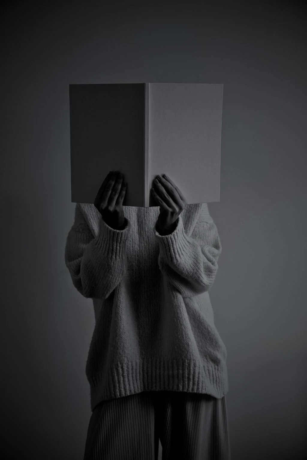 A person wearing a thick sweater holding a closed book in front of their face, obscuring their identity, with a neutral background.