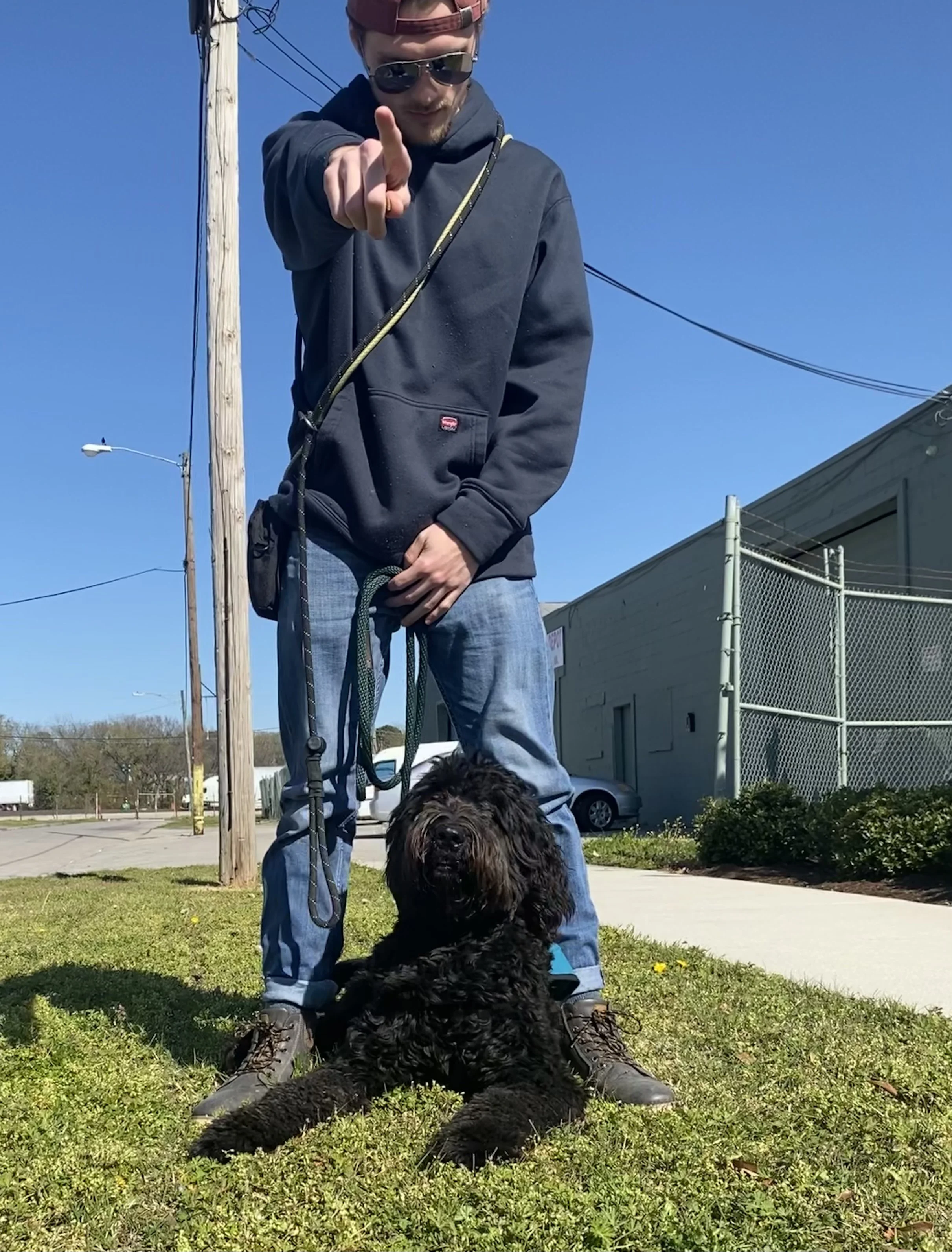 A man wearing sunglasses and a hoodie points towards the camera while standing on a grassy area next to a black dog sitting on the ground. The man has a leash attached to the dog and is wearing a backpack. There are power lines, a building, parked ca