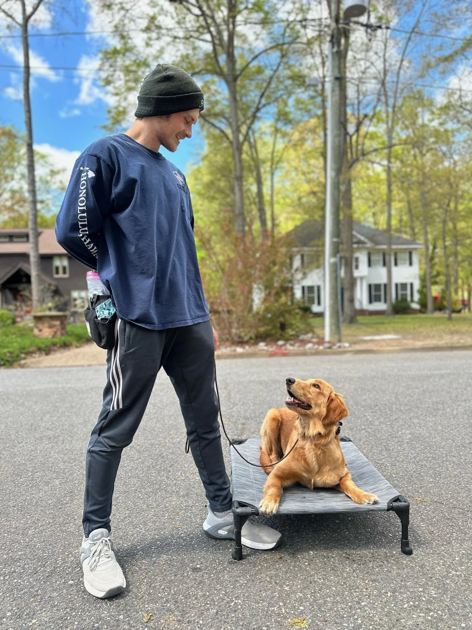 A man in a black beanie, navy long-sleeve shirt, and black athletic pants with white stripes stands next to a golden retriever puppy lying on a small elevated dog bed outdoors on a paved surface. The man is looking down at the puppy, who is looking u
