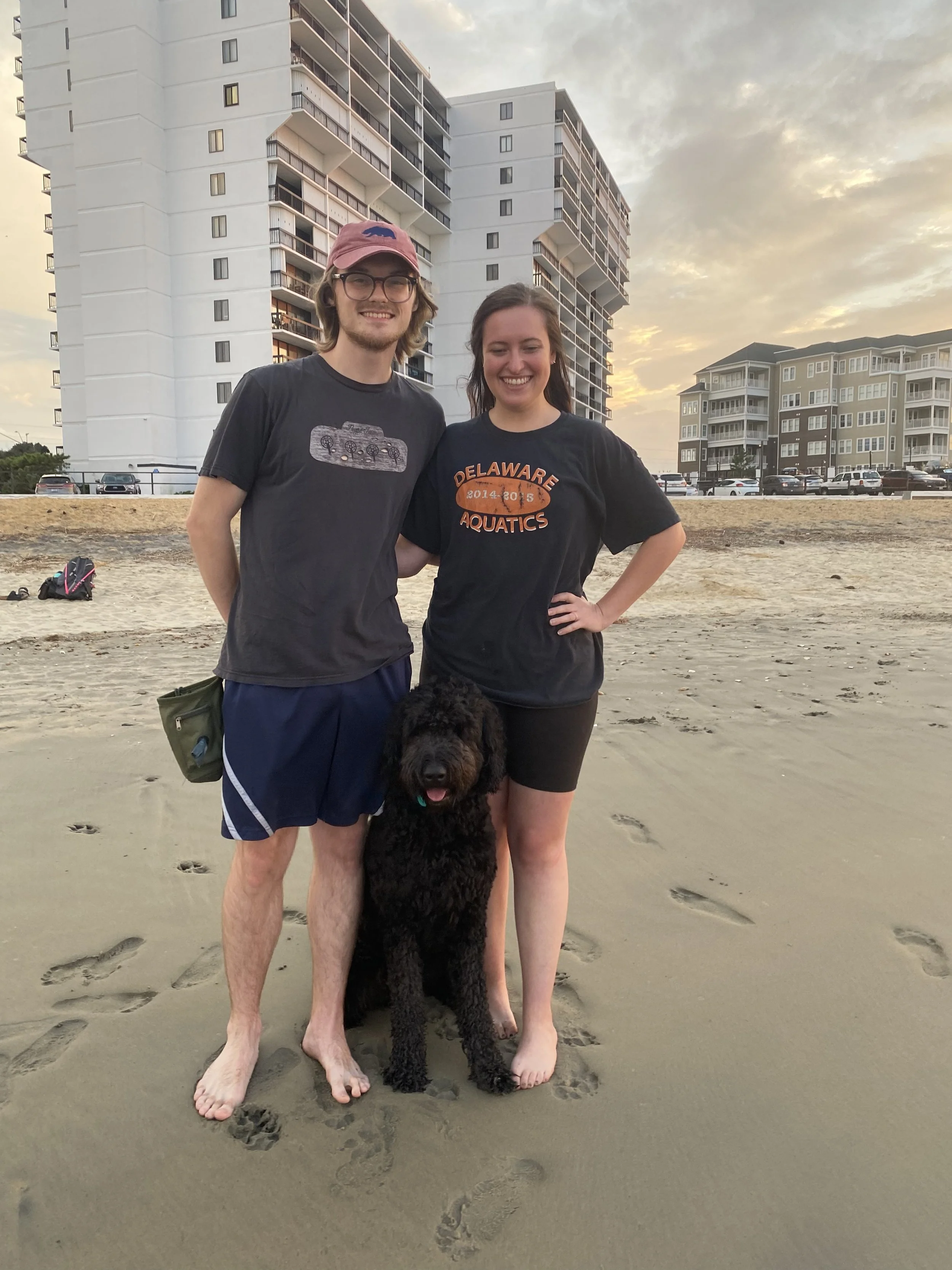 A smiling young man, woman, and a large black dog pose on a sandy beach during sunset with apartment buildings in the background.