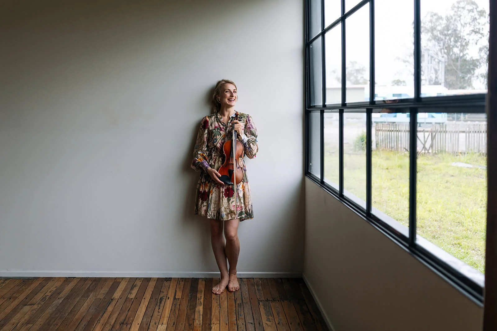 A woman holding a violin, standing barefoot in a room with wooden floor and a large window, smiling and looking to the side.