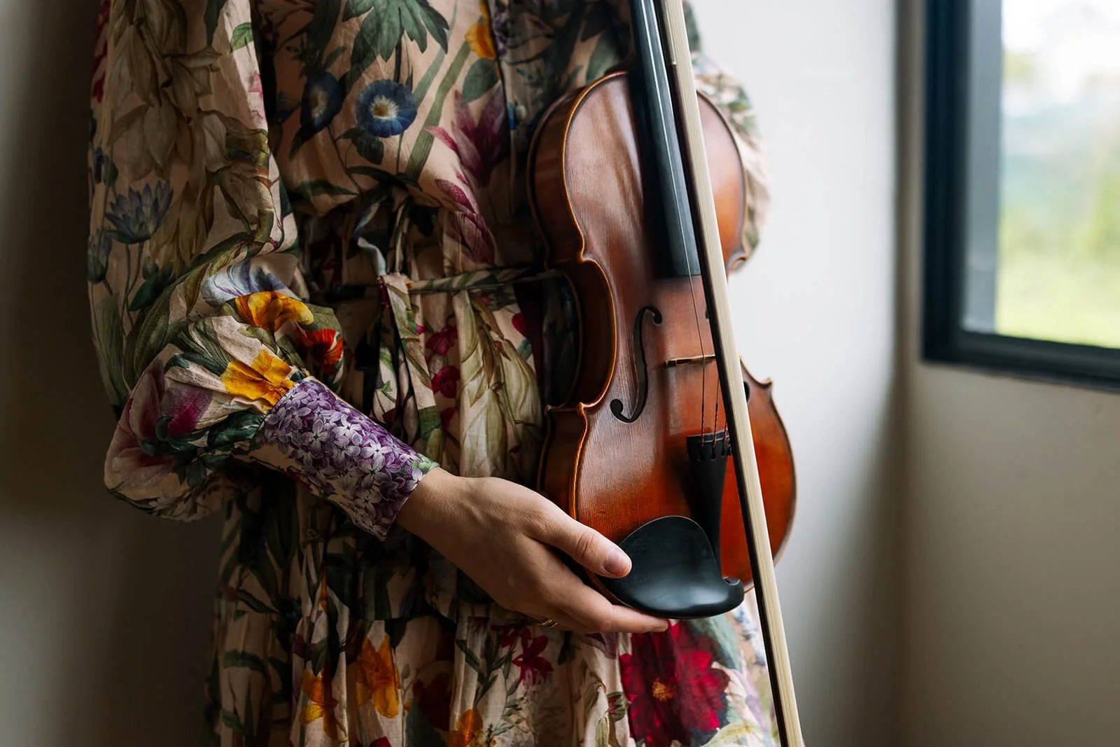 Tabea Sitte-Irwin person holding a violin and bow, wearing a floral patterned dress, standing near a window.