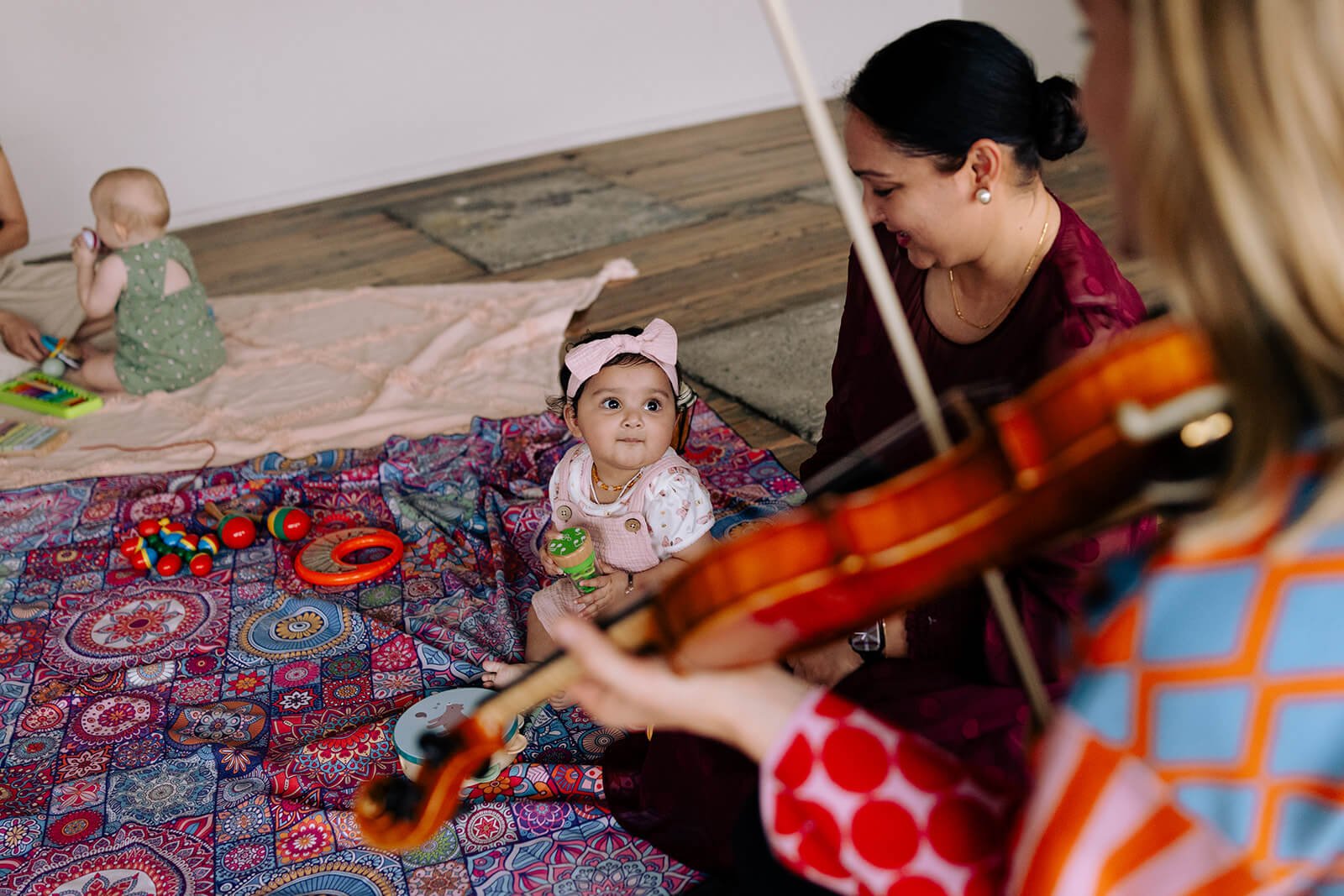 Mums and bubs music class at Klangfarben Creative Yeronga. Tabea playing a violin while a baby girl sits on a colorful blanket, looking up at her, surrounded by toys. A second child plays nearby on a pink blanket.