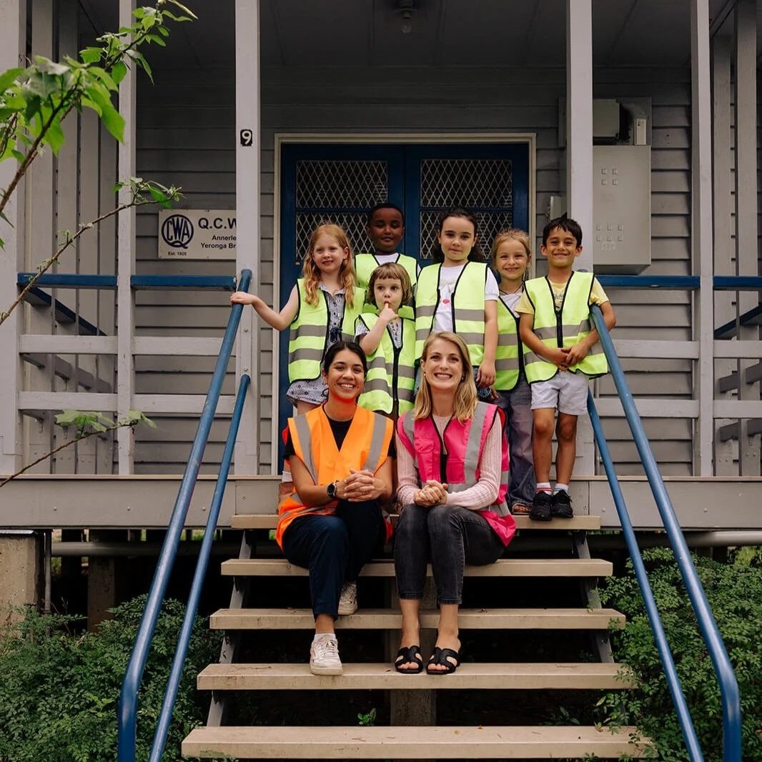 Group of children and two adult women sitting and standing on the steps of Klangfarben Creative, some children wearing yellow safety vests, and two women wearing orange and pink safety vests.