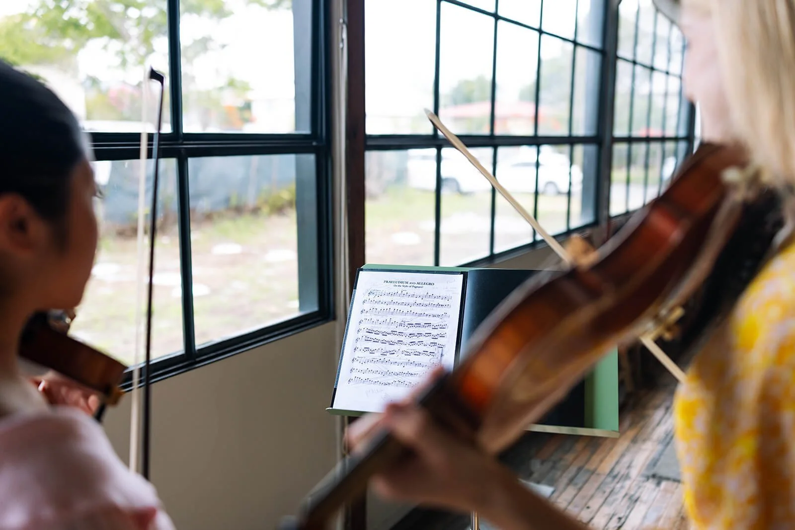 Two women playing violins and sheet music on a music stand in front of a window with a view of trees and a parking lot outside.