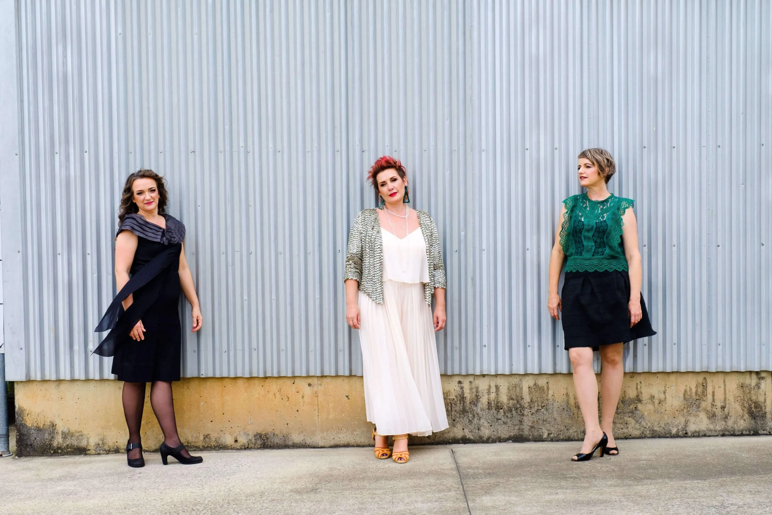 Muses Trio - Three women standing outdoors against a vertical metal wall, dressed in fashionable clothing.