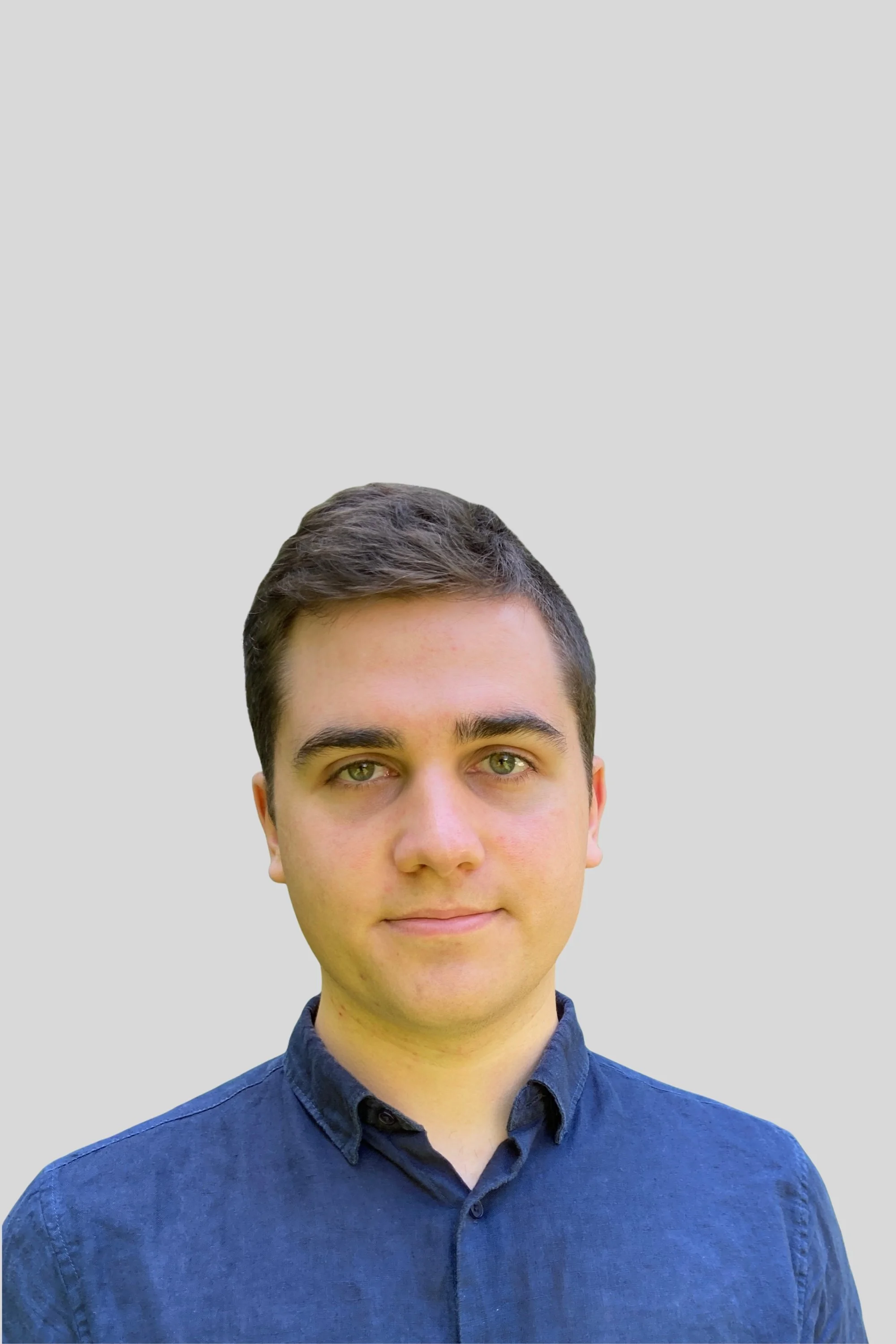 Portrait of piano teacher Nava, with short brown hair and light skin, wearing a dark blue button-up shirt, standing against a plain light gray background.