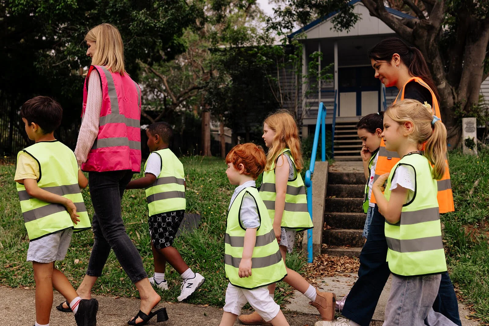 Young children and educators wearing bright yellow safety vests walking in line outside.