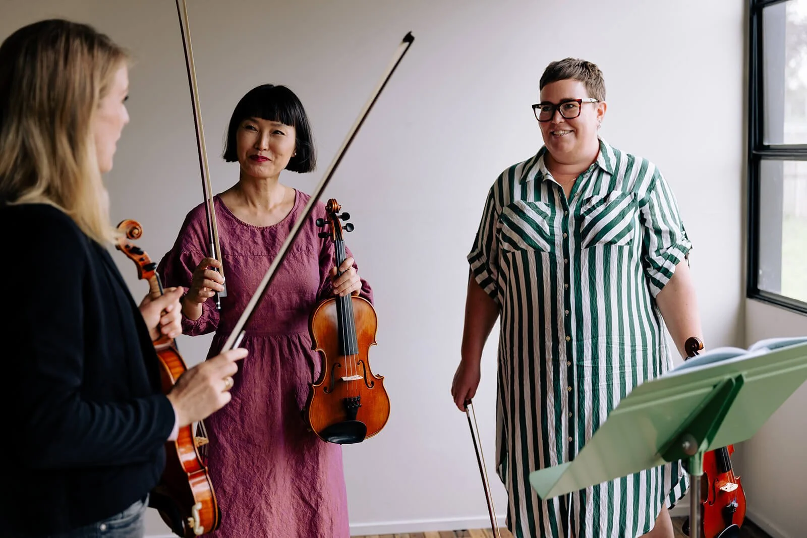 Three women in a music room holding violins and bows, with music stand near a window.