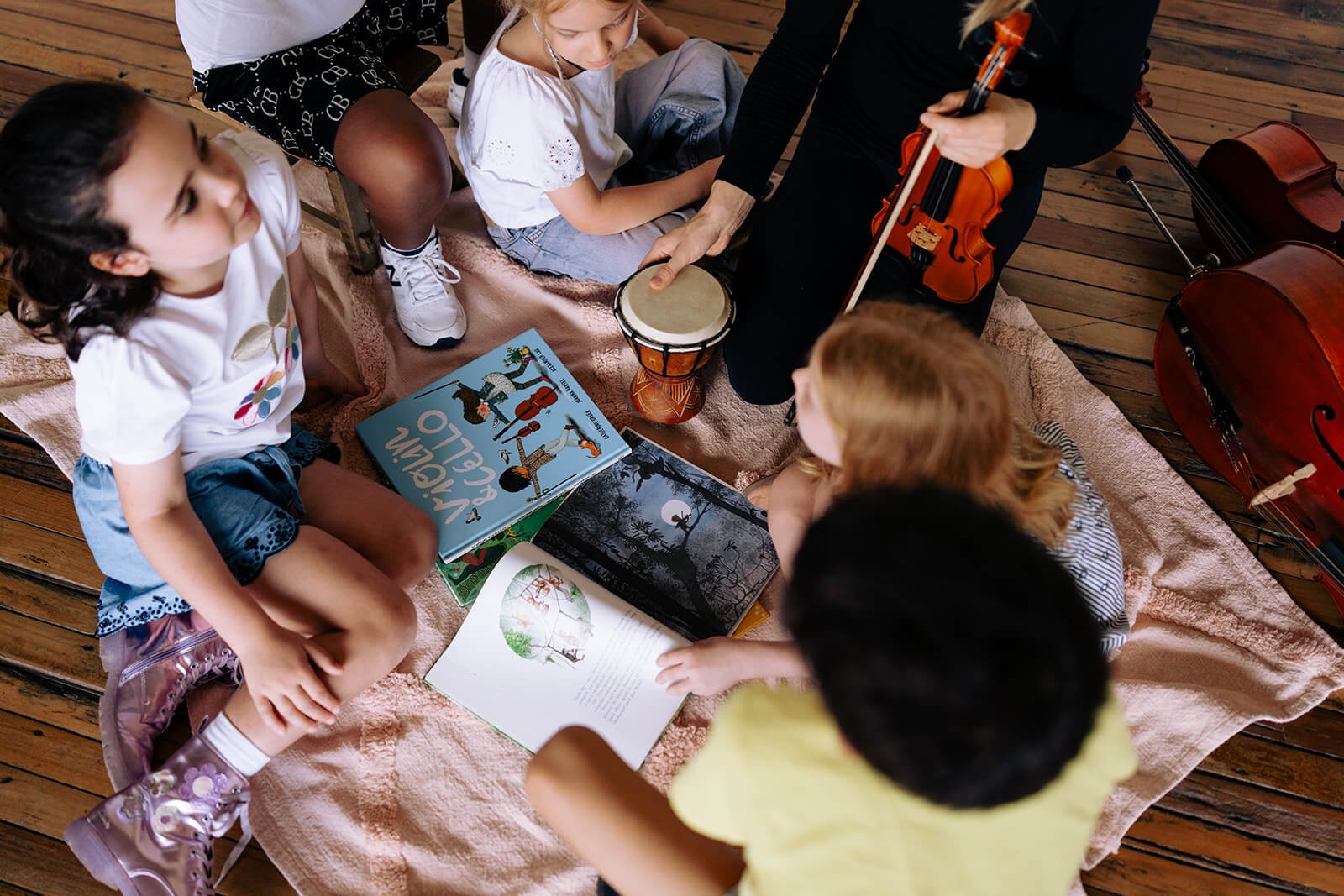 Children sitting on a blanket, surrounded by children's books and musical instruments, including a drum and a cello, in a cozy indoor setting.