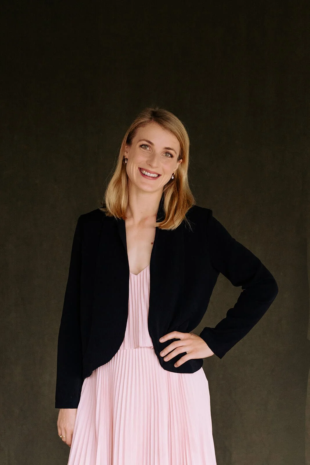 Tabea Sitte-Irwin with blonde hair, wearing a black blazer and a pink pleated dress, standing against a dark background, smiling and posing with one hand on her hip.