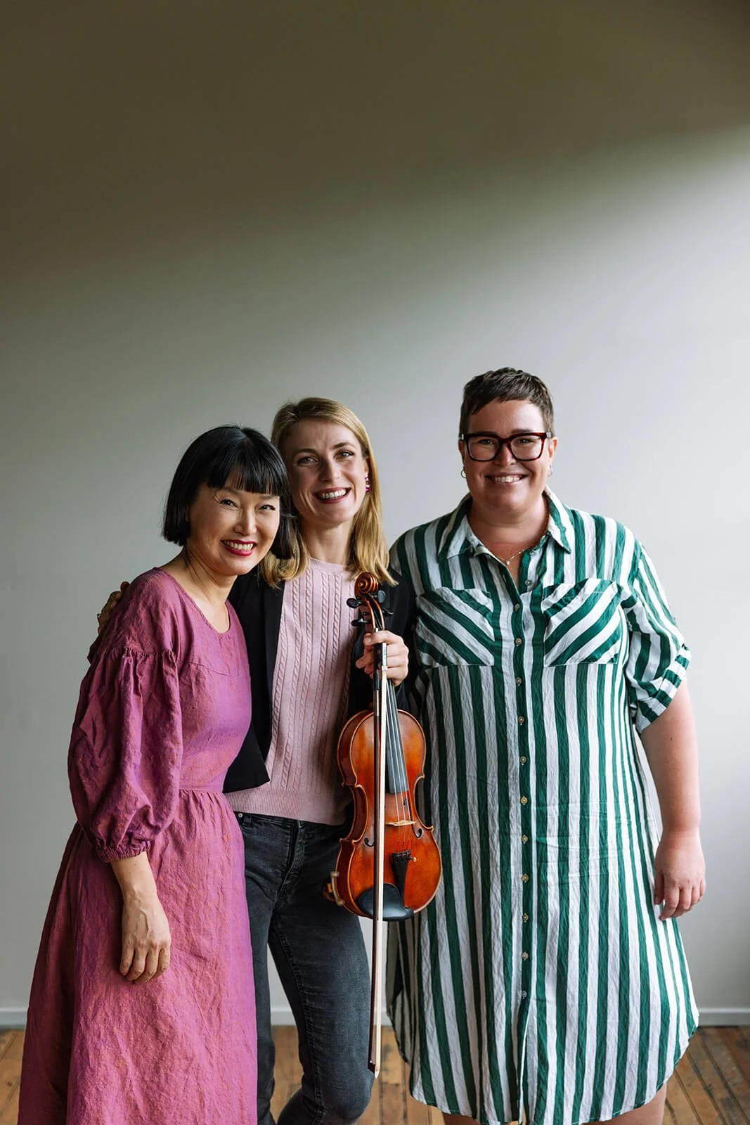 Three women standing together, one holding a violin, smiling at the camera in a bright room.