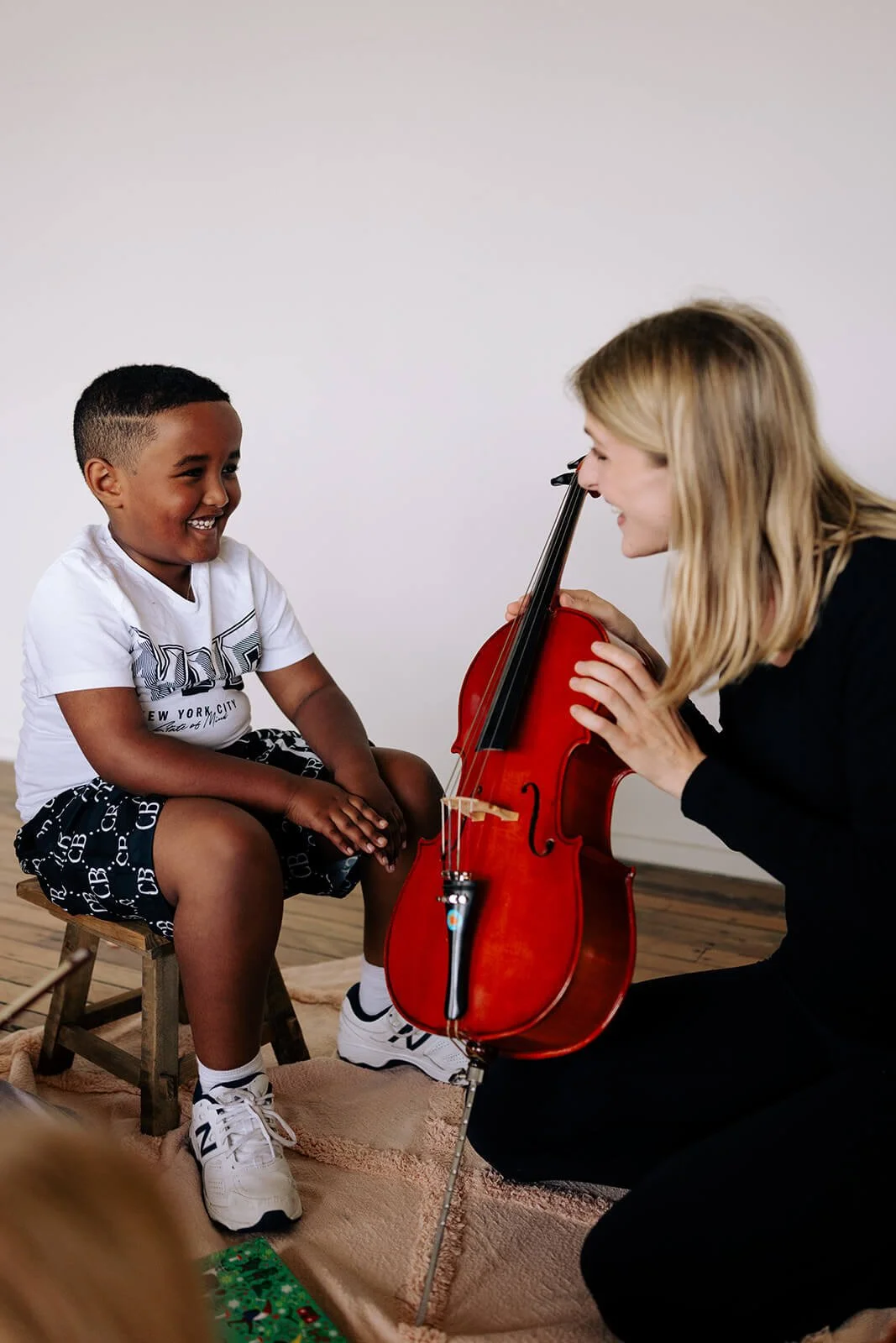 A boy sitting on a small stool, smiling, while his violin teacher Tabea Sitte-Irwiin shows him the violin.