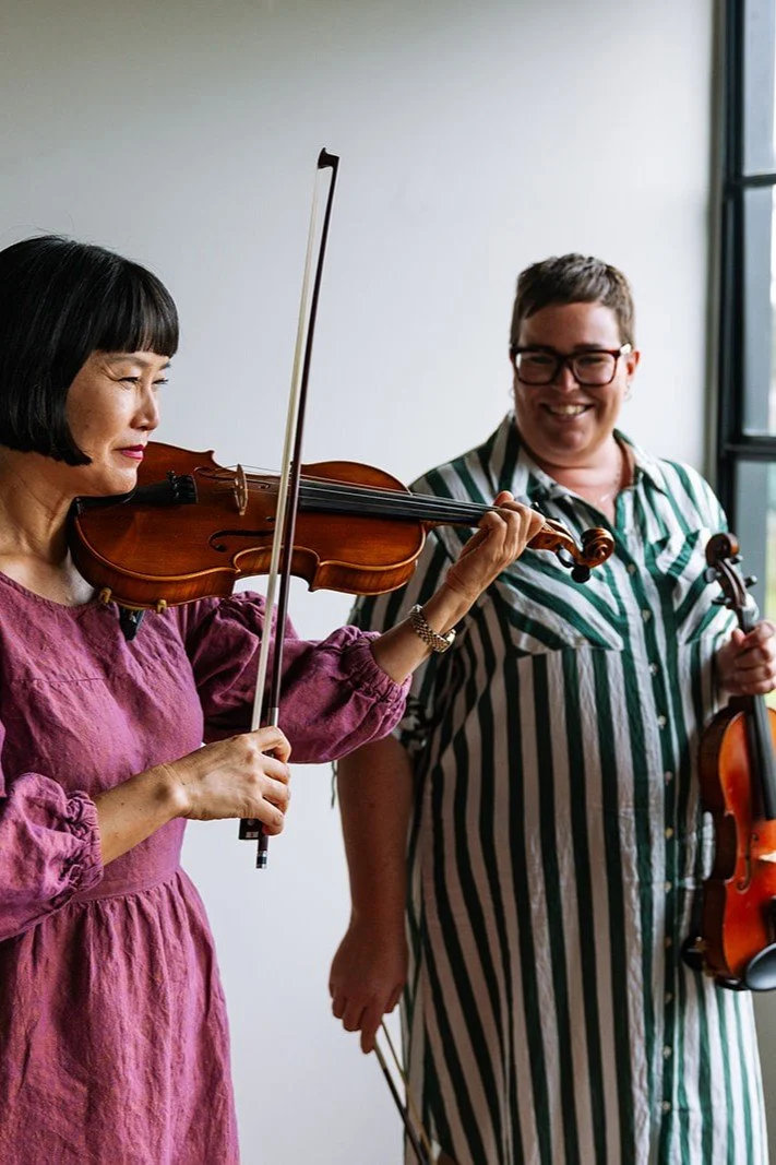 Two women, one playing the violin and the other holding a second violin, smiling and standing near a large window.