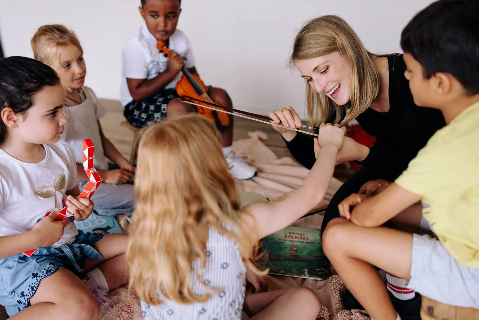 A woman and a young girl are playing a viola together while other children watch, sitting on a carpet.