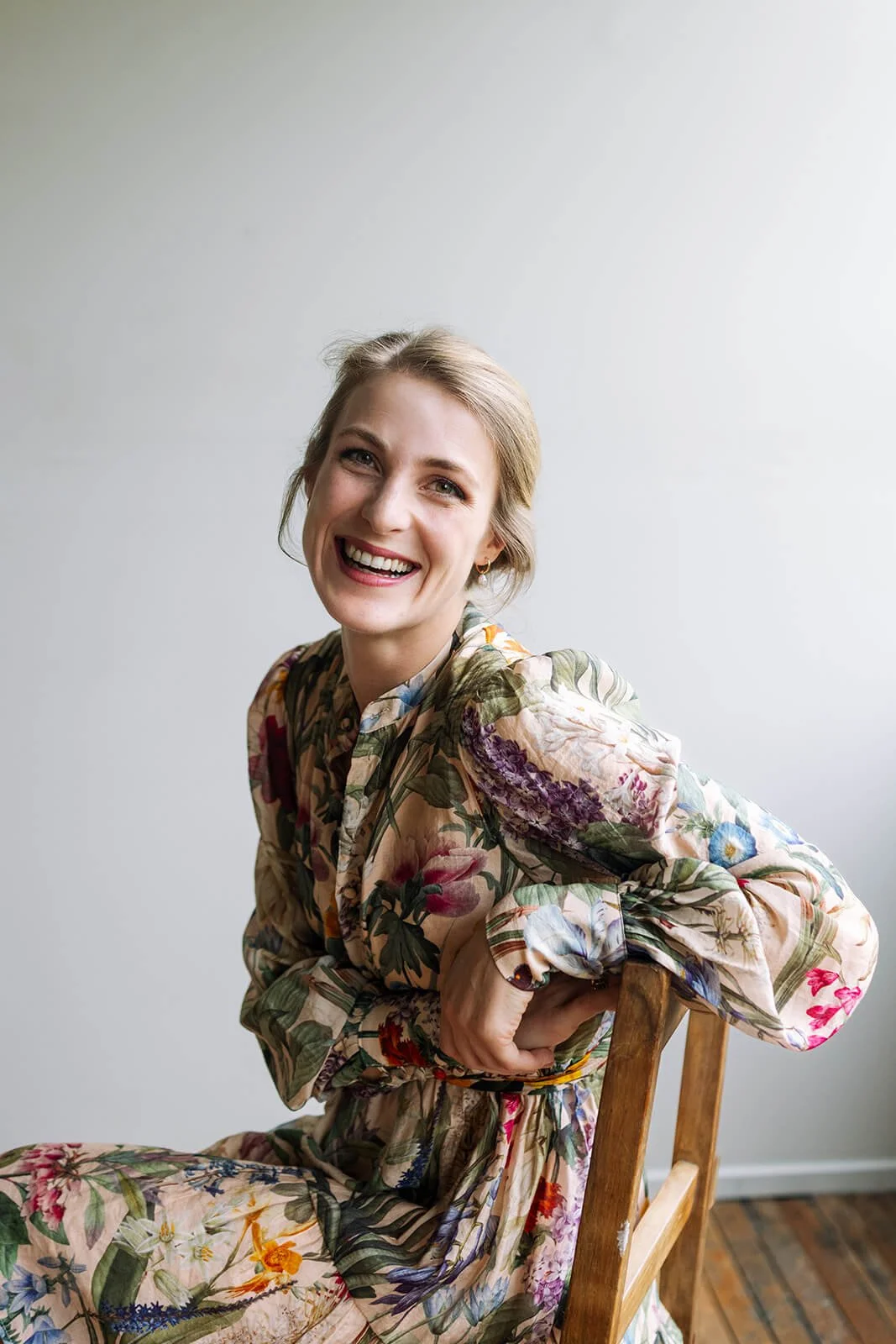 Violin teacher and concert violinist Tabea Sitte-Irwin, smiling with blond hair in a floral dress, sitting on a wooden chair against a plain wall.