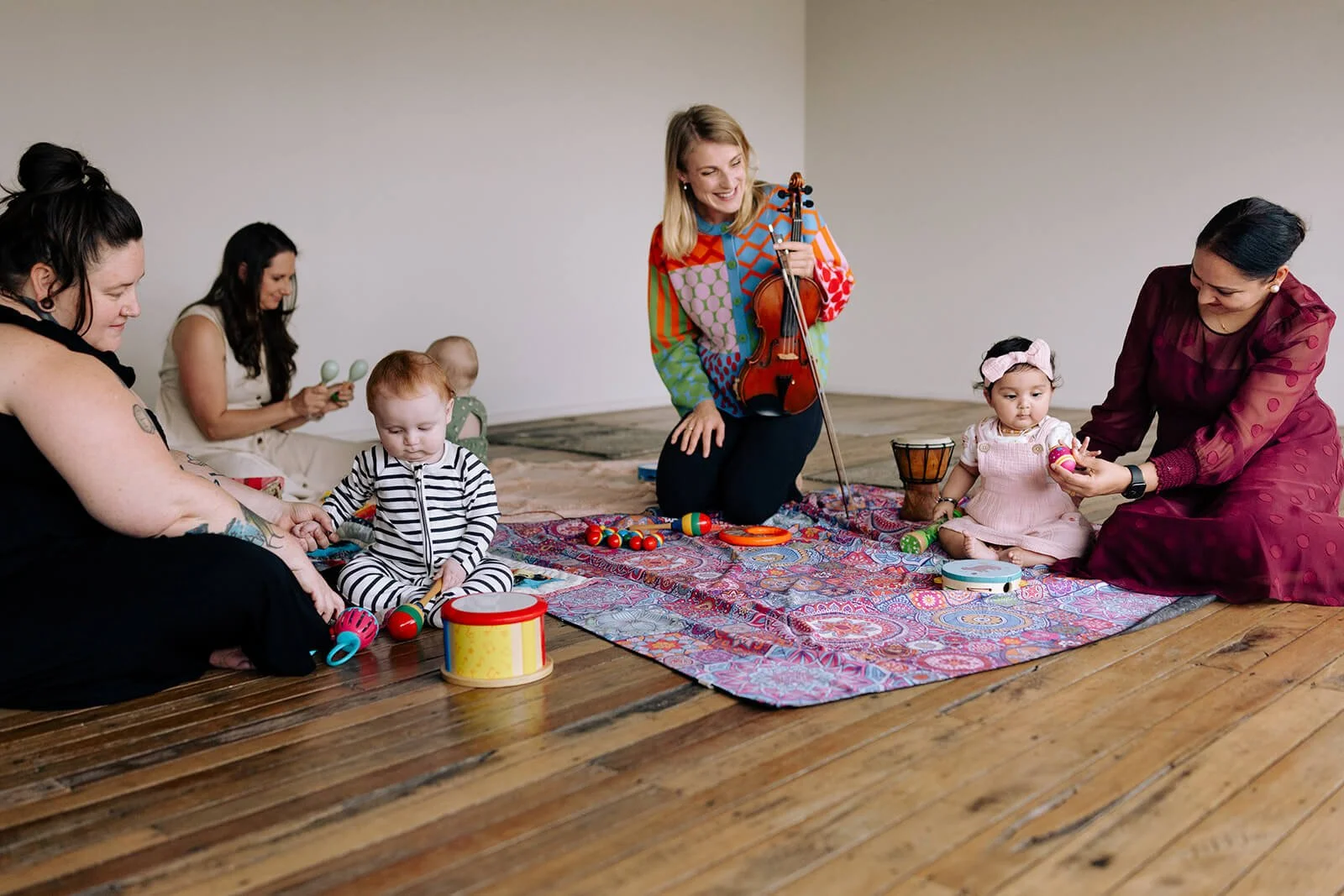 Women and children sitting on a colorful rug playing with toys, one woman playing the violin, in a room with wooden floor and blank wall.