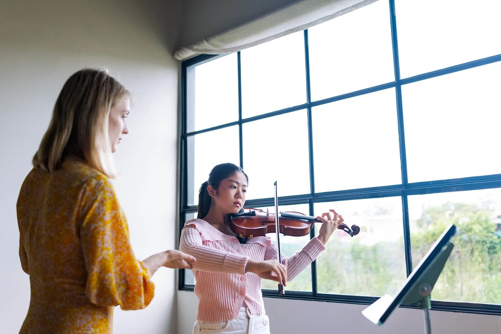 violin lessons, Yeronga - a young girl playing the violin while her instructor watches. They are in a room with a large window letting in daylight.