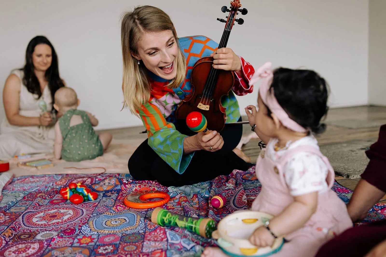 Babies and toddler music classes. Tabea Sitte-Irwin playing a violin and a maraca for a young girl with dark hair and a pink headband, sitting on a colorful blanket with toys.