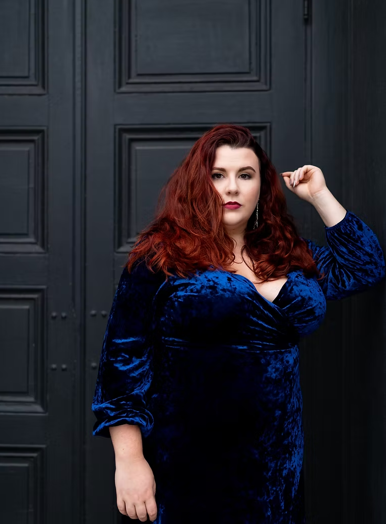A woman with long, wavy red hair, wearing a dark blue velvet dress, standing in front of a dark door with panels and rivets.