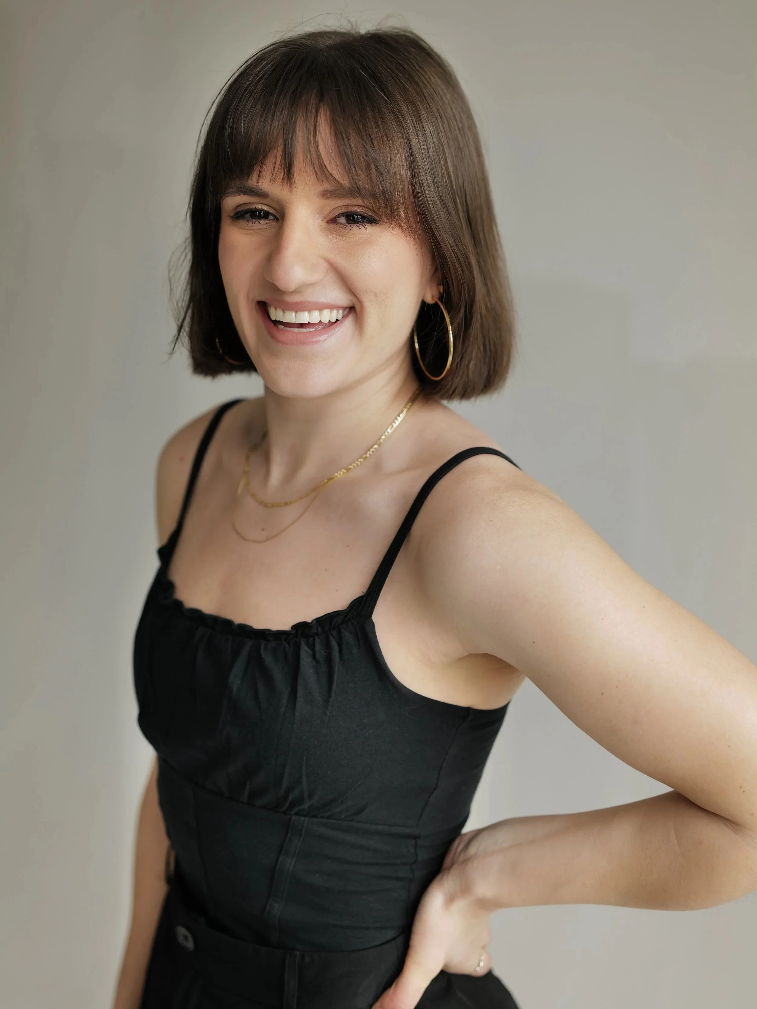A smiling woman with short brown hair, wearing a black sleeveless top, gold hoop earrings, and layered gold necklaces, posing with her hand on her hip against a neutral background.