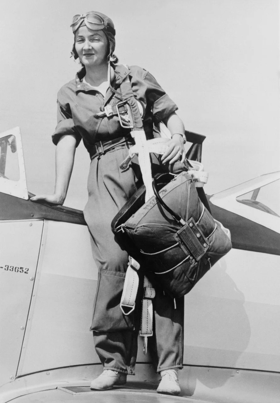 Black and white photograph of a woman standing on an airplane wing, wearing a pilot uniform, helmet with goggles, and carrying gear and a bag.