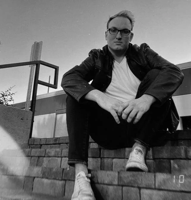 A black and white photo of a young man sitting on outdoor brick stairs, wearing glasses, a leather jacket, a t-shirt, and sneakers, with a modern building and a basketball hoop in the background.