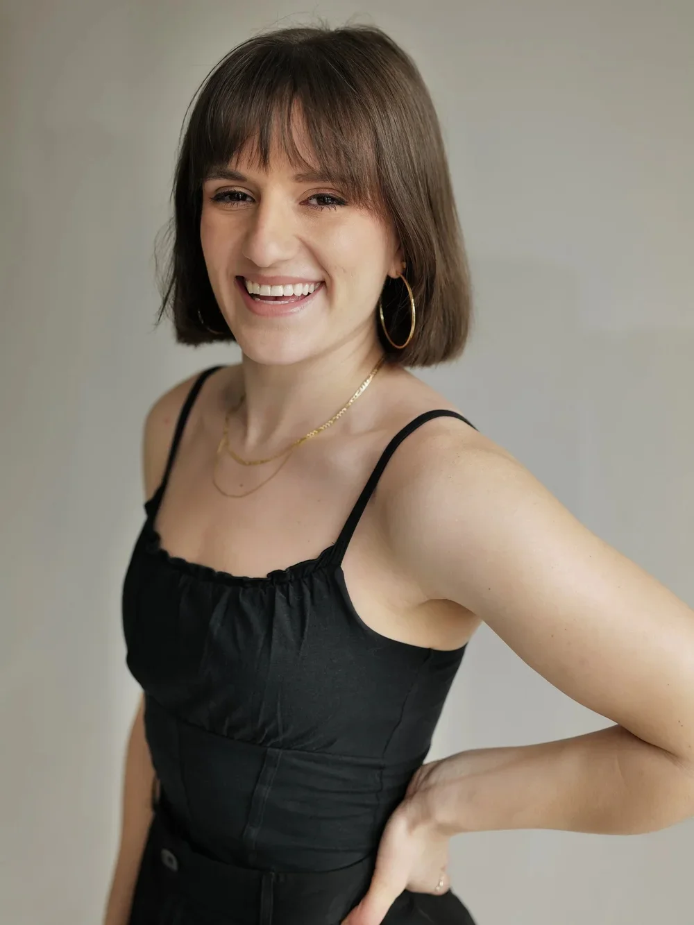A smiling woman with shoulder-length brown hair, wearing a black spaghetti strap top and gold jewelry, standing against a plain light-colored background.