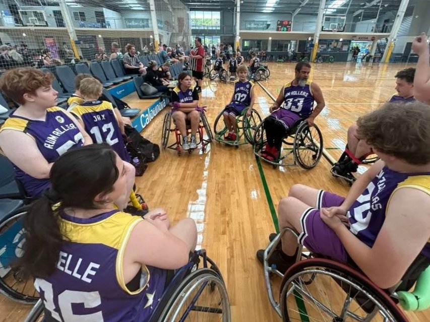 Wheelchair basketball players sitting on the court and listening to their coach during a game or practice in an indoor sports facility.
