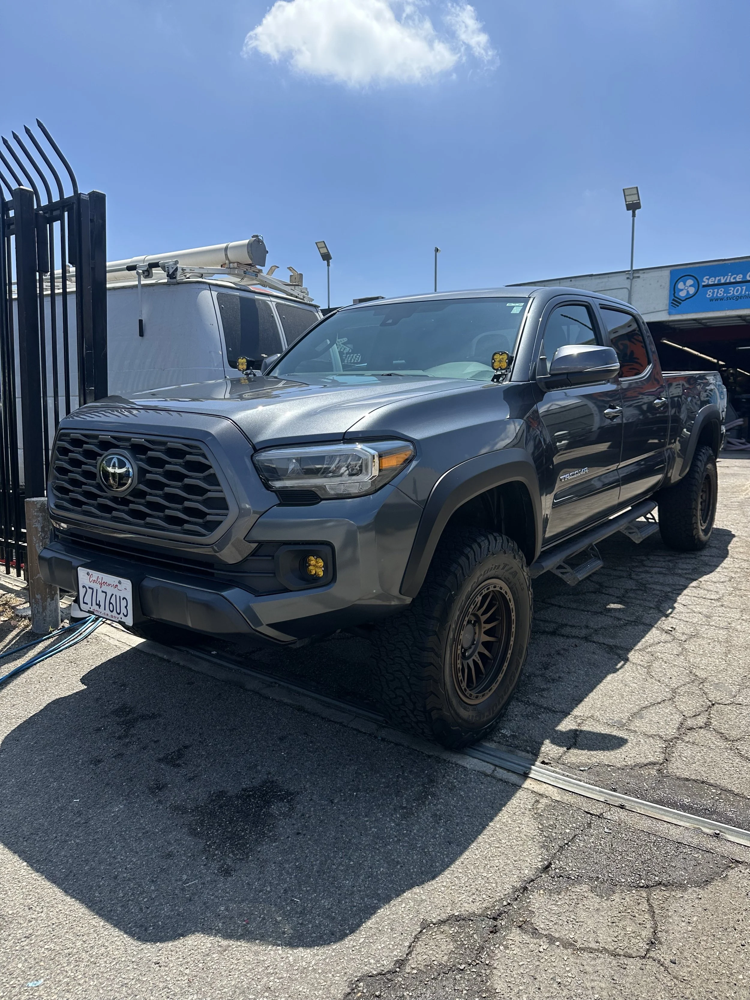 A new gray Toyota Tacoma pickup truck parked next to a white utility van at a commercial location, with a blue sky overhead and a parking lot with cracks and a metal barrier in front of the truck.