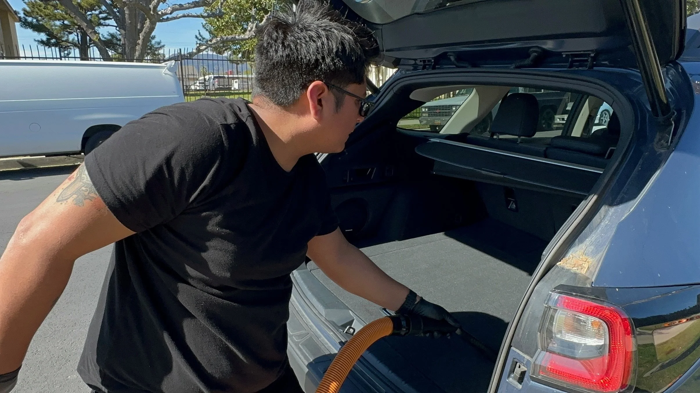 A man with black hair, sunglasses, and a tattoo on his arm is leaning into the open trunk of a gray car, possibly vacuuming or cleaning inside on a sunny day.