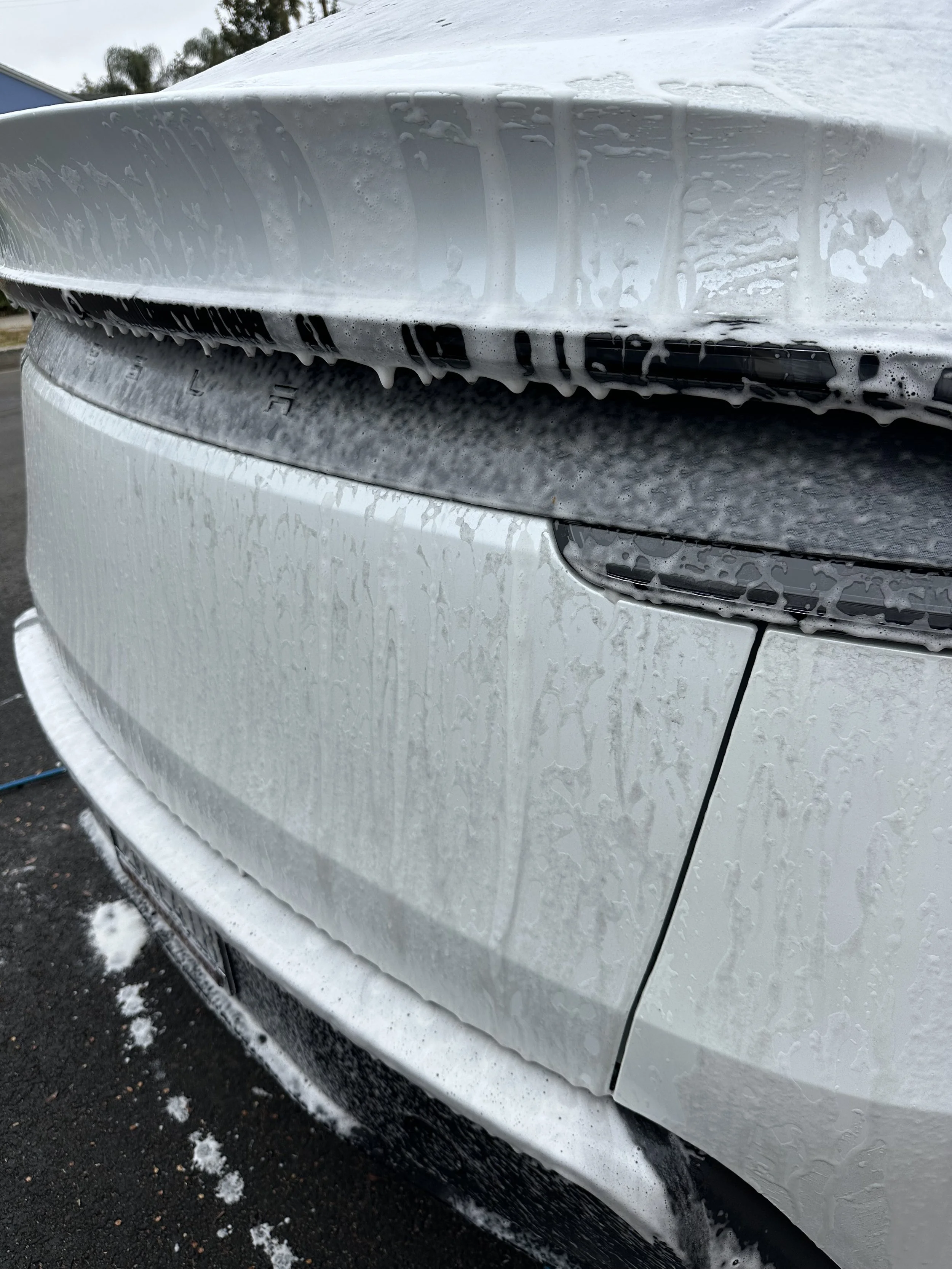 Close-up of a white car's rear end, with soap suds and water on the surface, the car is parked with some snow on the ground.