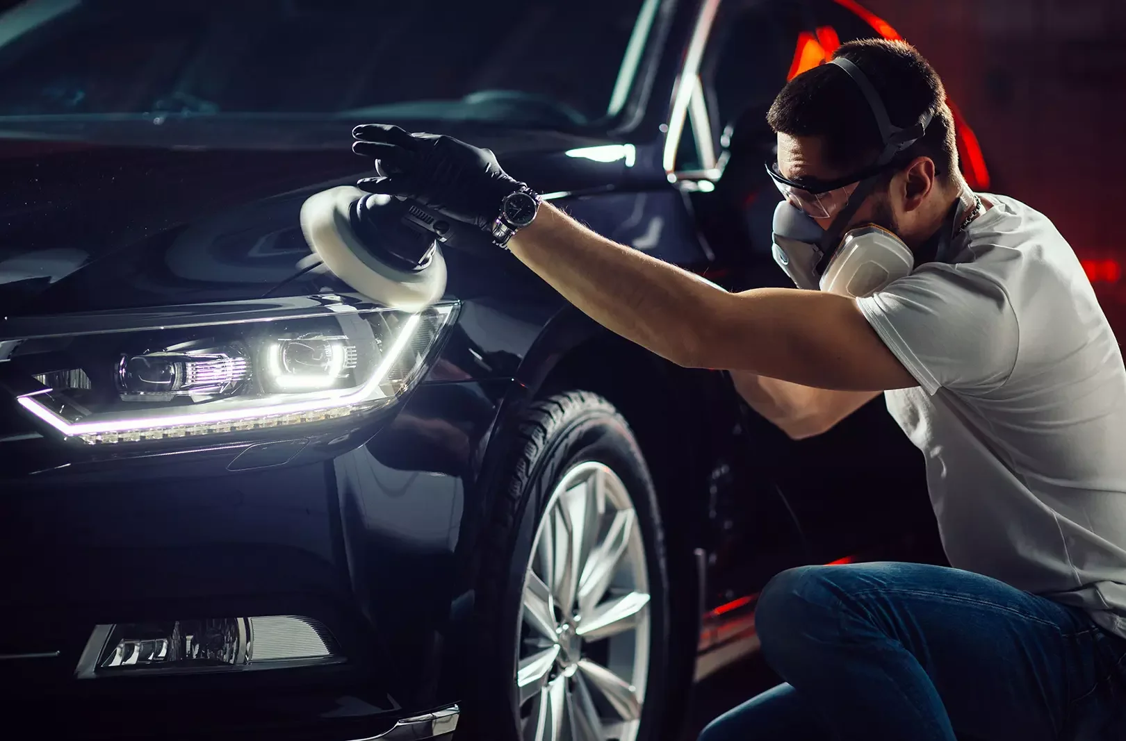 A man wearing gloves, a mask, goggles, and a watch is polishing the front headlight and fender of a black car with a polishing machine in a garage or workshop.