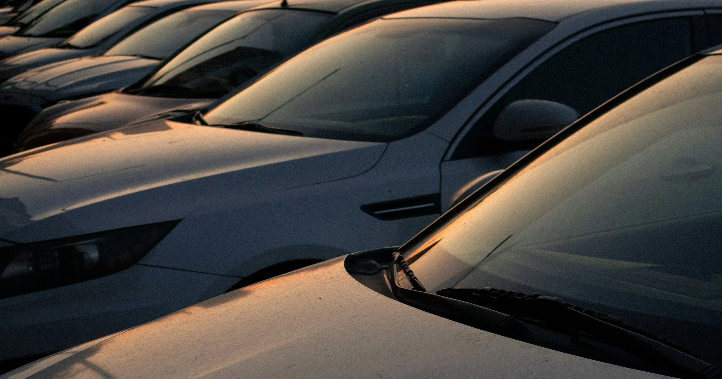 Multiple cars parked in a lot at sunset, with reflections of the setting sun on their surfaces.
