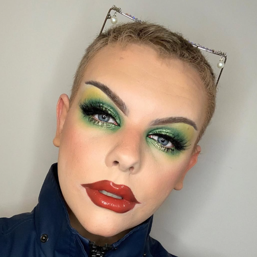 Person with short hair and vibrant green eye makeup, large false eyelashes, shiny red lipstick, wearing glasses with pearl accents on top, dressed in a dark jacket, against a plain background.