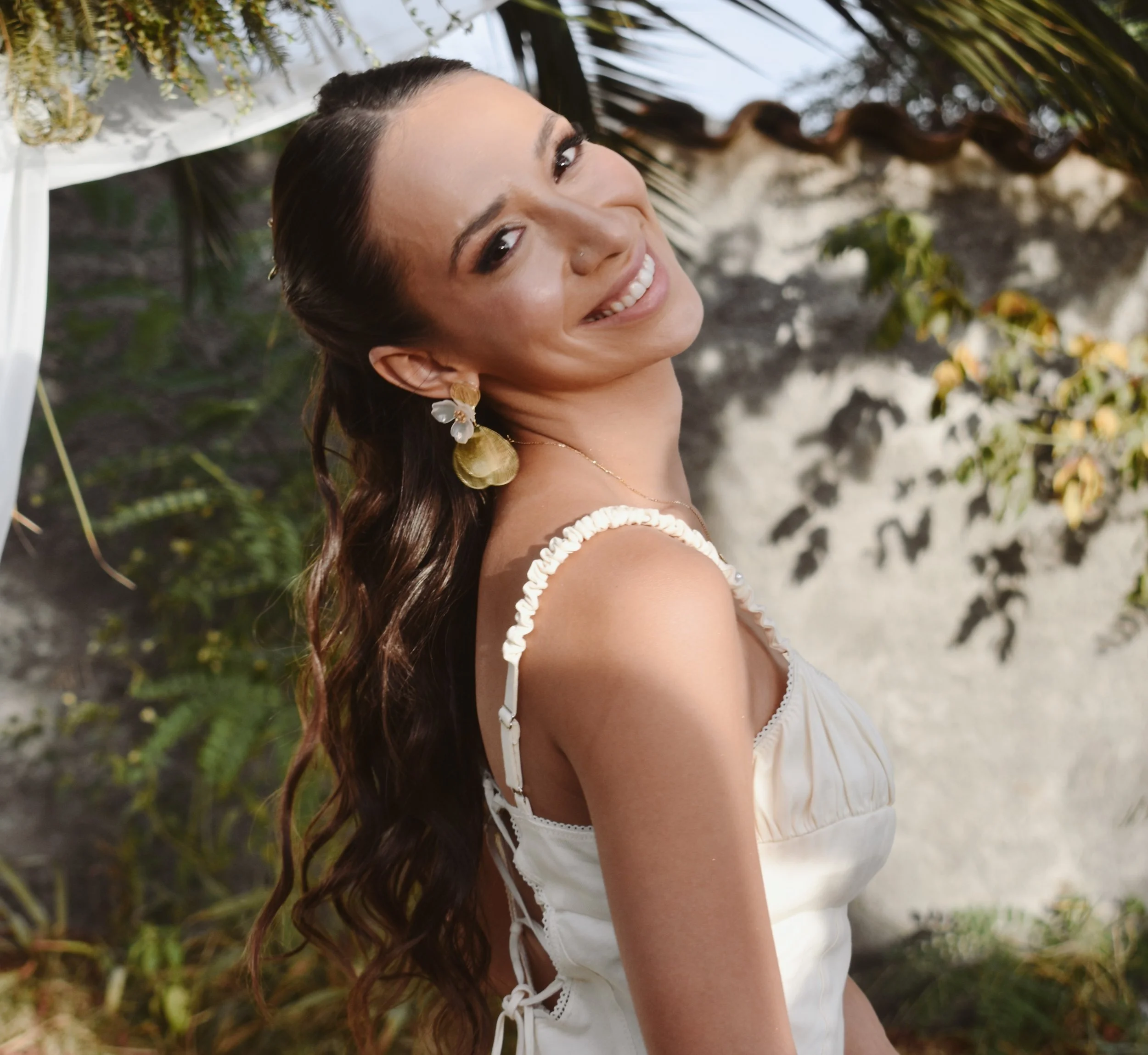 Une femme souriante portant une robe blanche, avec de longs cheveux bouclés, des boucles d'oreilles pendantes en or et en cristal, se tenait sous une structure décorative avec des plantes en arrière-plan, en plein air, probablement dans un jardin ou une station balnéaire.
