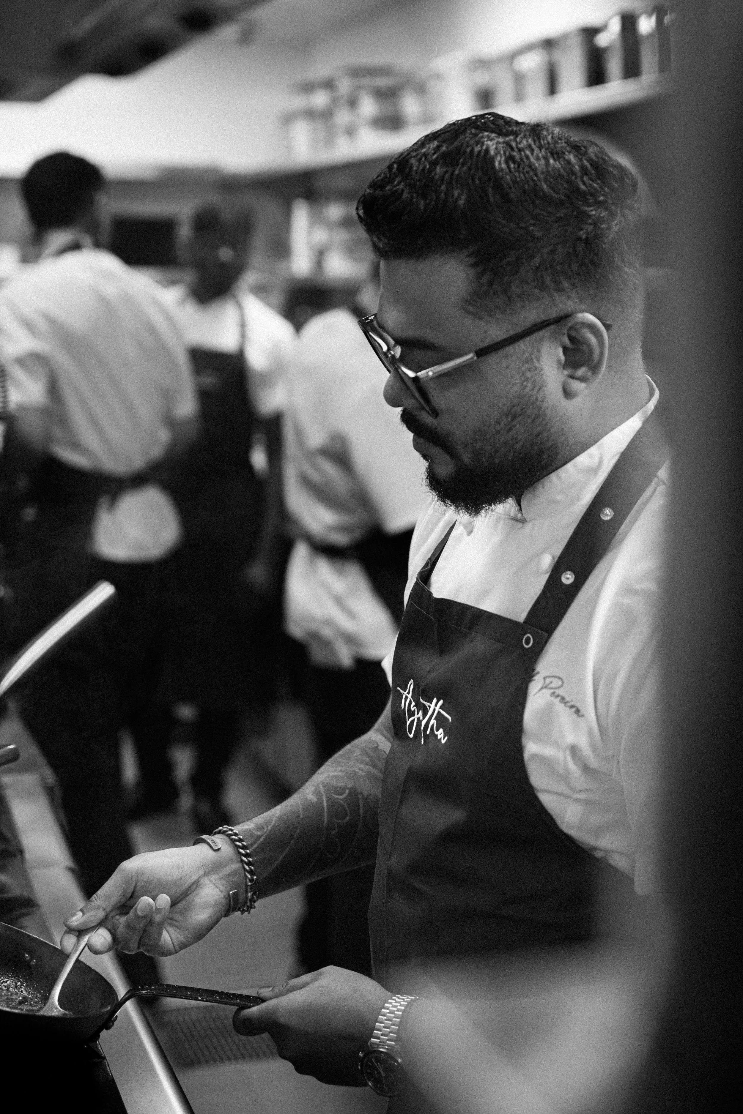 A man wearing glasses, a white shirt, and a black apron cooking in a kitchen with others in the background, in black and white.