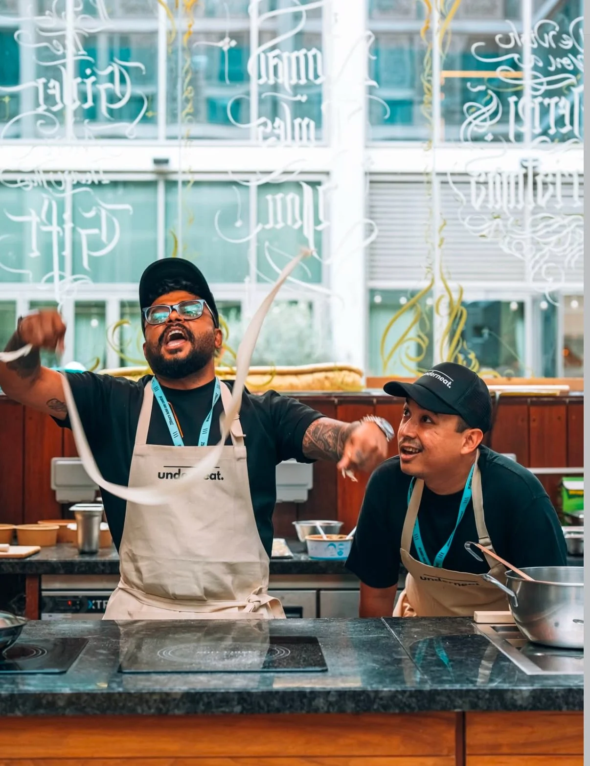 Two men in black shirts and aprons celebrating in a kitchen with a festive atmosphere, one wearing glasses and a cap, the other wearing a black cap, surrounded by kitchen appliances and utensils.